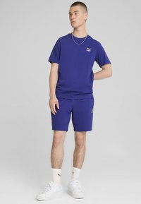 Modré sportovní tričko a odpovídající šortky, vyrobené z hladkého materiálu. Obsahuje malé bílé logo na hrudi a na boku šortek, doplněné bílými teniskami.