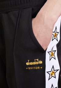 Schwarze Sporthosen mit einem seitlichen weißen Streifen, der goldene und schwarze Sternmuster aufweist. Goldenen Stickerei-Logo und den Text "EDITOR".