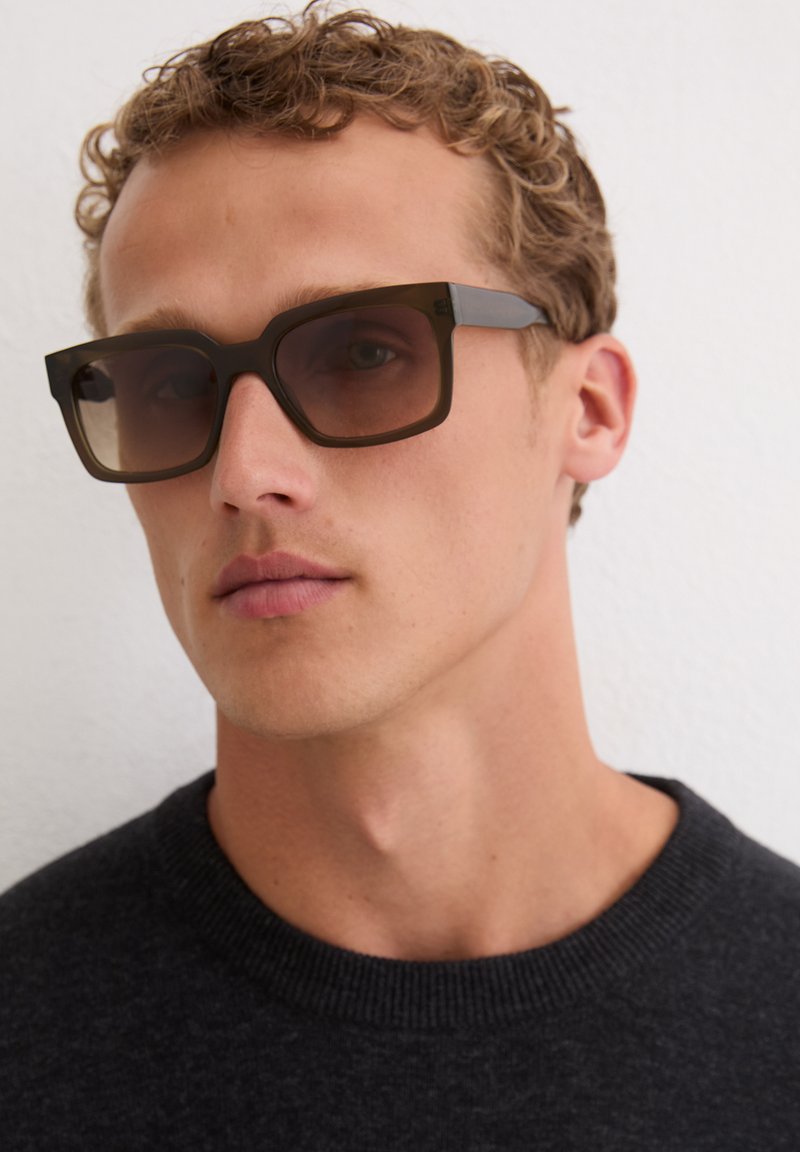 Lunettes de soleil rectangulaires marron avec des verres dégradés ; monture épaisse mate ; portées avec un pull en maille foncé ; fond neutre.