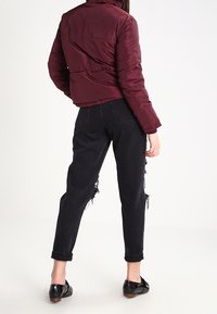 Chaqueta de plumas burdeos con cuello alto, cierre de cremallera y un diseño recortado, combinada con unos vaqueros negros desgastados y zapatos negros brillantes.