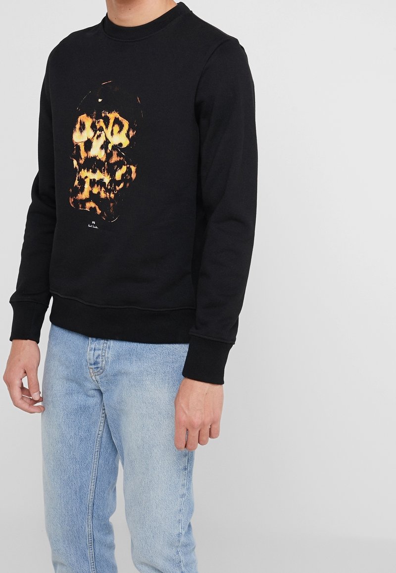 Homme portant un sweat-shirt noir avec un design abstrait de crâne orange et jaune, associé à un jean bleu clair, debout devant un fond uni.