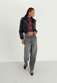 Woman with braided hair wearing a black cropped leather jacket, red patterned crop top, gray high-waist jeans, and black pointed heels walking.