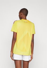 Camisola desportiva amarela com um padrão texturizado, mangas curtas e gola redonda. Apresenta detalhes em gradiente e um painel em malha nas costas.