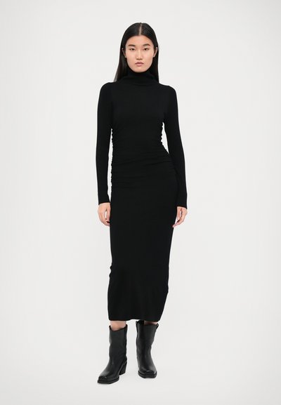 Fekete bordázott kötött ruha magas nyakkal, hosszú ujjakkal és testhez simuló sziluettel. A modell fekete bokacsizmával viseli. Simaságot sugárzó anyag.