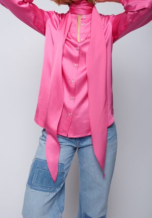 Person iført en lyserød satinbluse med fastgjort langt tørklæde og lyseblå jeans med stor denimlap på låret.