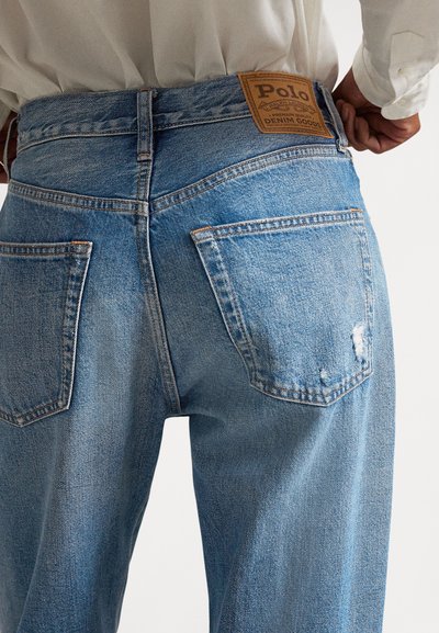 Inimese lähedalt võetud foto, kes kannab siniseid teksaseid, millel on tagataskud ja vöökohal pruun Polo Ralph Laureni silt.