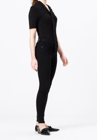 Musta lyhythihainen paita, jossa on kaulus, yhdistetty istuviin mustiin farkkuihin. Täydennetty kiiltävillä mustilla loafereilla, joissa on teksturoitu pinta.