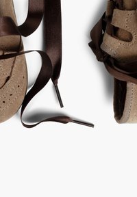 Los zapatos de gamuza marrón presentan una puntera redondeada y un intrincado detalle perforado, con largas cintas de satén marrón chocolate para el cordón.