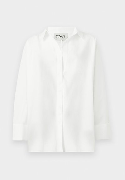 Bílá košile s dlouhými rukávy, zapínáním na knoflíky, límečkem a širokými manžetami, uvnitř límce označená nápisem "TOVE", zobrazena na jednobarevném pozadí.