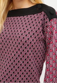 Zwart en roze gebreide trui met een geometrisch patroon, voorzien van een bootnek en zwarte knoopaccenten op de schouder.
