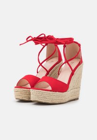 Sandali con zeppa in suede rosso, con lacci alla caviglia, punte aperte e tacchi in stile espadrilles intrecciati. Il design presenta cinturini incrociati e texture lisce.