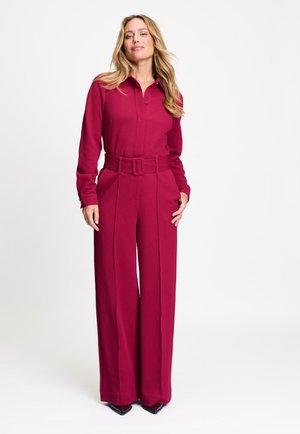Vrouw staand, gekleed in een magenta jumpsuit met lange mouwen, een brede riem en wijde pijpen tegen een effen witte achtergrond.