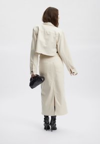 Kobieta w kremowej krótkiej kurtce i dopasowanej długiej spódnicy, trzymająca czarną kopertówkę, ubraną w czarne wysokie botki, na białym tle.