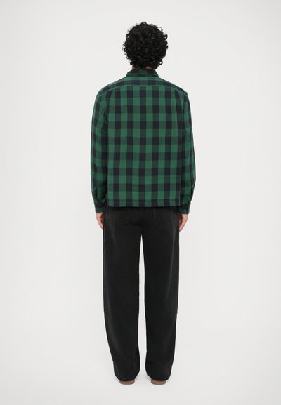 Camisa quadriculada verde e preta, de mangas compridas, com colarinho, de corte reto. Usada com calças pretas largas, apresentando um ajuste descontraído.