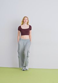 Top curto castanho com mangas curtas, combinado com calças cargo cinzas claras com bolsos laterais, usando ténis cinzentos, em pé sobre um chão verde.