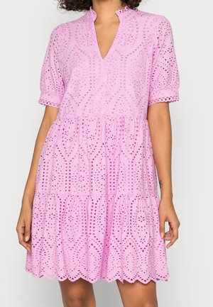 Nainen, joka pukeutunut lilaan, kirjailtua silmäreikämekkoon, jossa on pussihihat ja aaltoileva helma, seisoo yksinkertaista taustaa vasten.