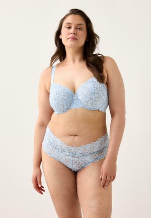 Kvinde med langt mørkt hår iført lyseblå blonde-bh og matchende højtaljede blondeunderbukser, stående mod en ensfarvet lys baggrund.