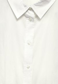 Camisa de cuello blanco hecha de tela suave, con cierre frontal de botones y botones de plástico transparente. Diseño minimalista con costuras bien acabadas.