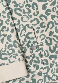 Tissu beige avec imprimé léopard vert, montrant un poignet de manche et une petite poche avec un bouton-pression rond en argent.