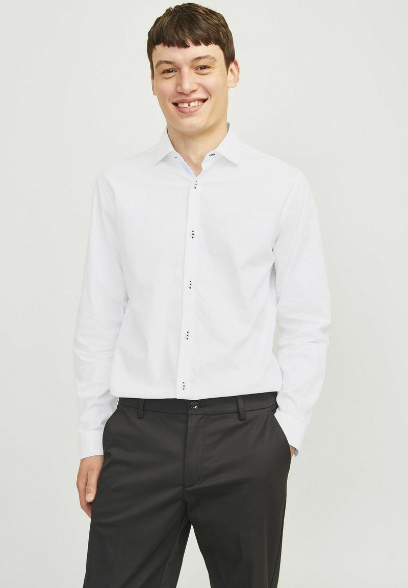 Chemise blanche à manches longues avec col boutonné, présentant des boutons noirs sur le devant. Associée avec un pantalon sombre, à texture minimale.