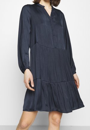Femme portant une robe à volants bleu foncé, manches longues, longueur genou, avec un col en V et des détails froncés au niveau du tissu.