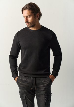 Hombre con barba y cabello de longitud media, vistiendo un suéter negro de manga larga y pantalones cargo negros, con las manos en los bolsillos mirando hacia abajo.