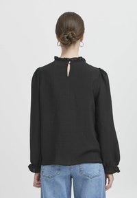 Blusa nera a maniche lunghe con scollo arricciato e dettaglio a chiave sul retro. Tessuto morbido e testurizzato, maniche arricciate e polsini aderenti.