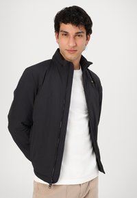 Black lightweight jacket with a high collar, zip closure, and minimal design. Worn over a white t-shirt, featuring a small brand tag.