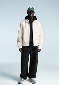 White zip-up jacket over a black hoodie, paired with loose black pants and gray sneakers. The model wears a green cap with a white logo.