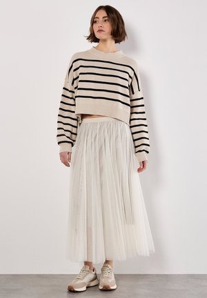 Femme portant un pull cropped beige avec des rayures noires, une longue jupe en tulle blanche et des baskets beiges, debout contre un mur blanc.
