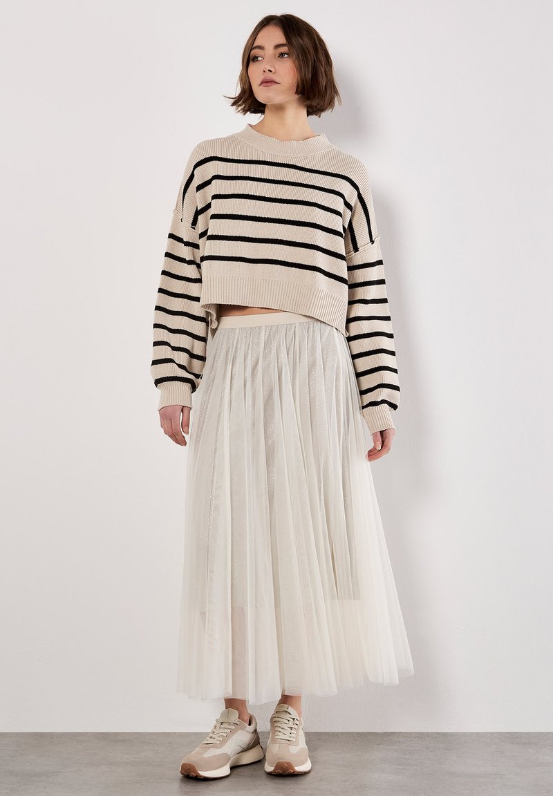 Femme portant un pull cropped beige avec des rayures noires, une longue jupe en tulle blanche et des baskets beiges, debout contre un mur blanc.