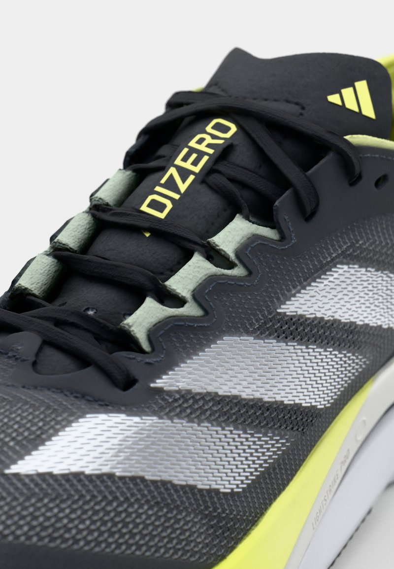 Czarne sportowe buty z odblaskowymi srebrnymi paskami, jasnoszarymi akcentami, teksturowaną siatkową cholewką i czarnymi sznurowadłami. Etykieta "Zero" w kolorze żółtym na języku.