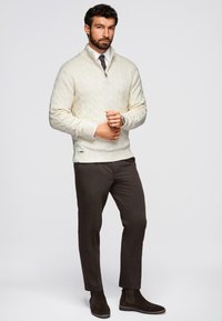 Mężczyzna noszący kremowy sweter z splotem, zapinany na zamek, białą koszulę, ciemny krawat, brązowe spodnie i ciemne zamszowe buty, stojący na jednolitym tle.