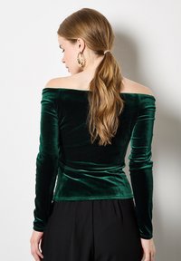 Grünes Samt-Oberteil mit schulterfreiem Design, langen Ärmeln und taillierter Silhouette, das eine glatte Textur und einen subtilen Glanz aufweist.