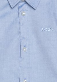 Camicetta azzurra chiara con bottoni, realizzata in tessuto liscio, con colletto a punta, bottoni bianchi e un logo ricamato "BOSS" sul petto.