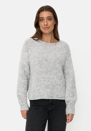 Vrouw met steil bruin haar, gekleed in een lichtgrijze grofgebreide trui en zwarte broek tegen een effen lichte achtergrond.