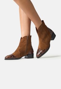 Picioare îmbrăcate cu botine din suede maro, cu accente decorative din piele texturată de culoare maro închis pe vârf și călcâi, și un toc mic.