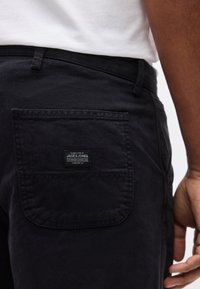 Κοντινό πλάνο ενός ατόμου που φοράει μαύρο παντελόνι με πίσω τσέπη που φέρει την ένδειξη "Jack & Jones" και ένα λευκό πουκάμισο που είναι μπηγμένο μέσα.