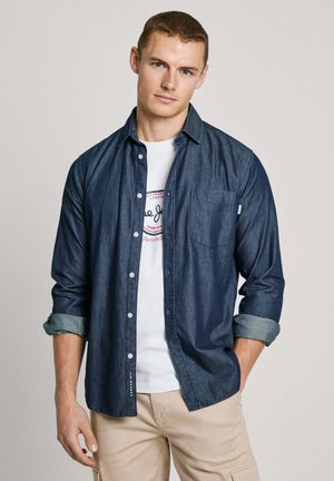 Dark blue denim button-up shirt with rolled sleeves, featuring a front pocket and light blue accents. Worn over a white graphic tee.