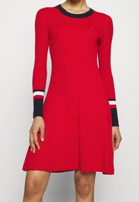 Punainen ribbineulemekko pitkillä hihoilla, jossa musta pyöreä pääntie ja raidoitusta hihansuissa punaisella, valkoisella ja mustalla.