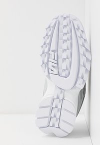 Semelle blanche de sneaker Fila avec motif de bande de roulement texturé et logo, positionnée verticalement contre des murs blancs.