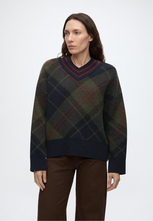 Femme portant un pull à col en V à carreaux verts et bleu marine avec des rayures rouges et un pantalon marron, debout devant un fond blanc uni.