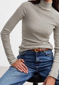 Top de cuello alto a rayas en crema y negro. Diseño ajustado con mangas largas, combinado con jeans azul de tiro alto y un cinturón marrón.