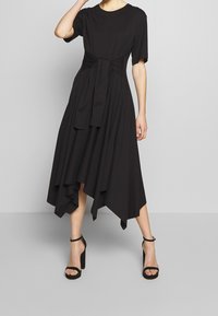 Kvinna klädd i en svart asymmetrisk midiklänning med korta ärmar och midjeband, parat med svarta högklackade sandaletter med ankelsnor.