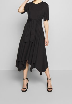 Mujer vistiendo un vestido midi negro asimétrico con mangas cortas y un lazo en la cintura, combinado con sandalias negras de tacón alto y tiras al tobillo.