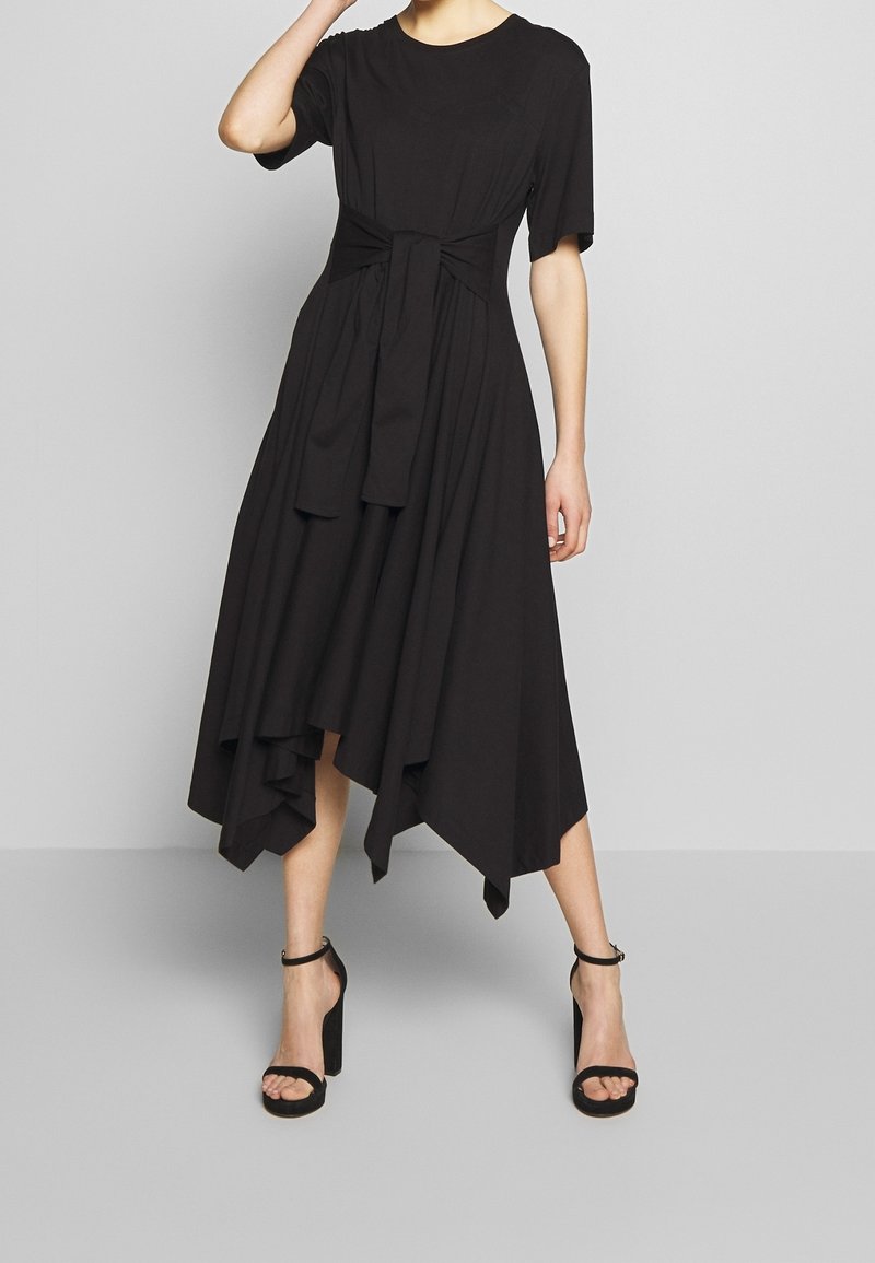 Kvinna klädd i en svart asymmetrisk midiklänning med korta ärmar och midjeband, parat med svarta högklackade sandaletter med ankelsnor.