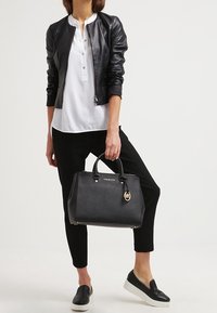 Donna che indossa una giacca di pelle nera, una blusa bianca, pantaloni neri e scarpe slip-on nere, mentre tiene in mano una borsa nera Michael Kors.