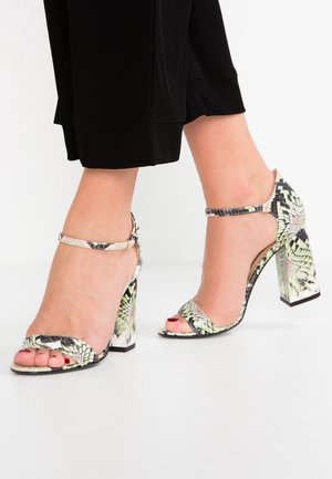 Voeten in sandalen met open tenen, blokhakken en enkelbandjes, met een lichtgroen en zwart slangenhuidpatroon, gecombineerd met zwarte cropped broek.