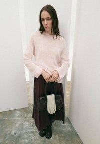 Suéter de punto rosa claro, falda plisada de color púrpura oscuro, botas negras hasta la rodilla, sosteniendo un bolso oscuro con guantes blancos. Superficies texturizadas.