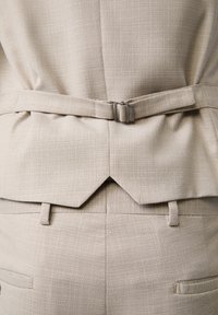 Beige Textile-Blazer mit strukturiertem Muster, seitlichem Schnallen-Detail und Schlitz auf der Rückseite, kombiniert mit passenden Hosen.